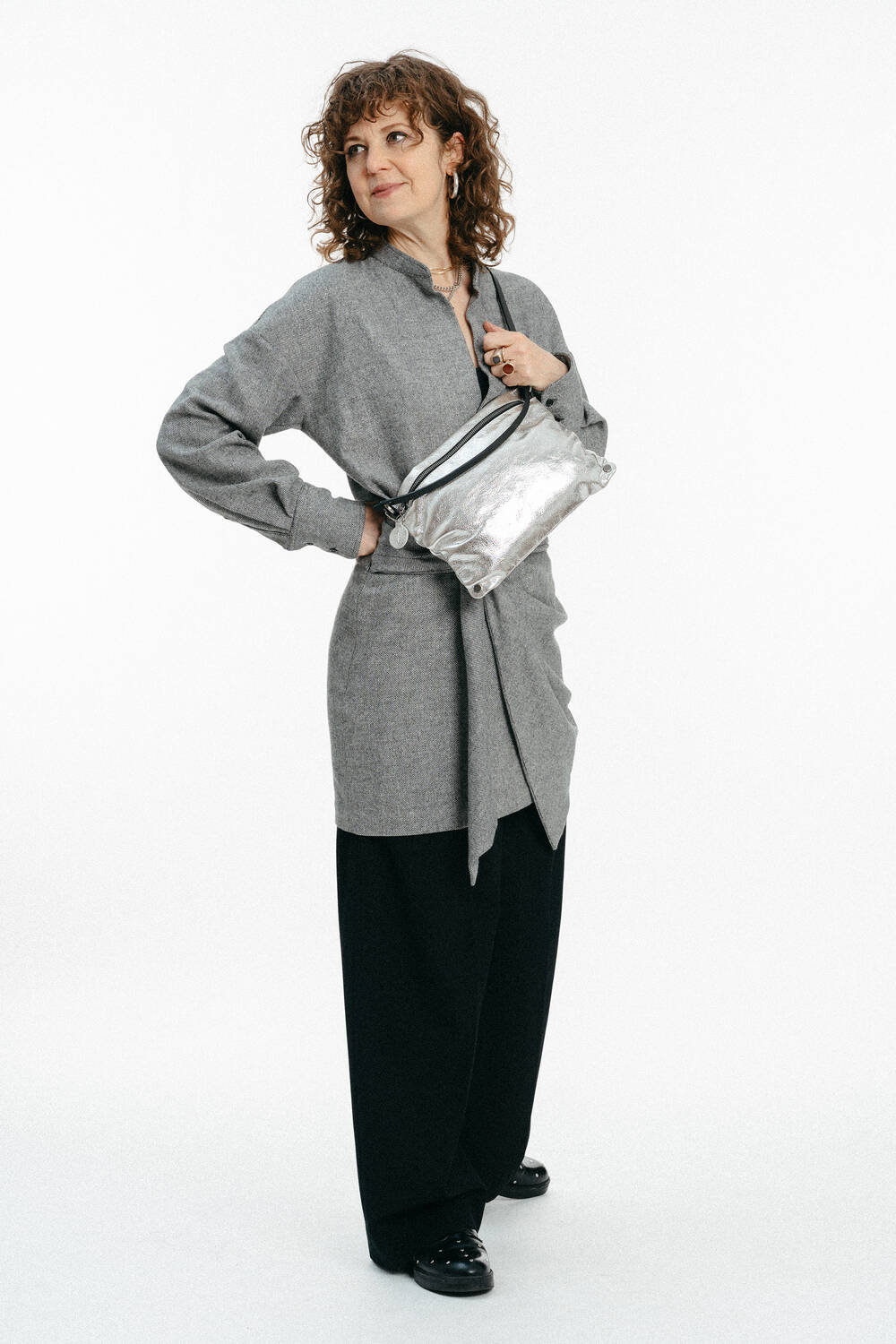 Frau in einer grauen Tunika und einer schwarzen Hose mit weitem Bein, gestylt mit einer silbernen INA KENT Metallic-Crossbody-Tasche und schwarzen Schuhen vor einem weißen Hintergrund.