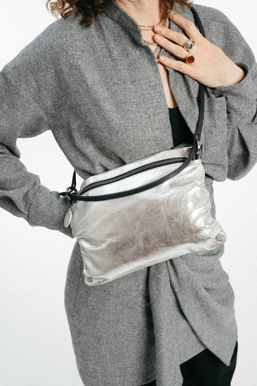 Person, die einen grauen Mantel mit Gürtel und ein schwarzes Oberteil trägt, eine silberne INA KENT-Metallic-Crossbody-Tasche in der Hand hält und mit der linken Hand Ringe und eine Halskette zeigt.