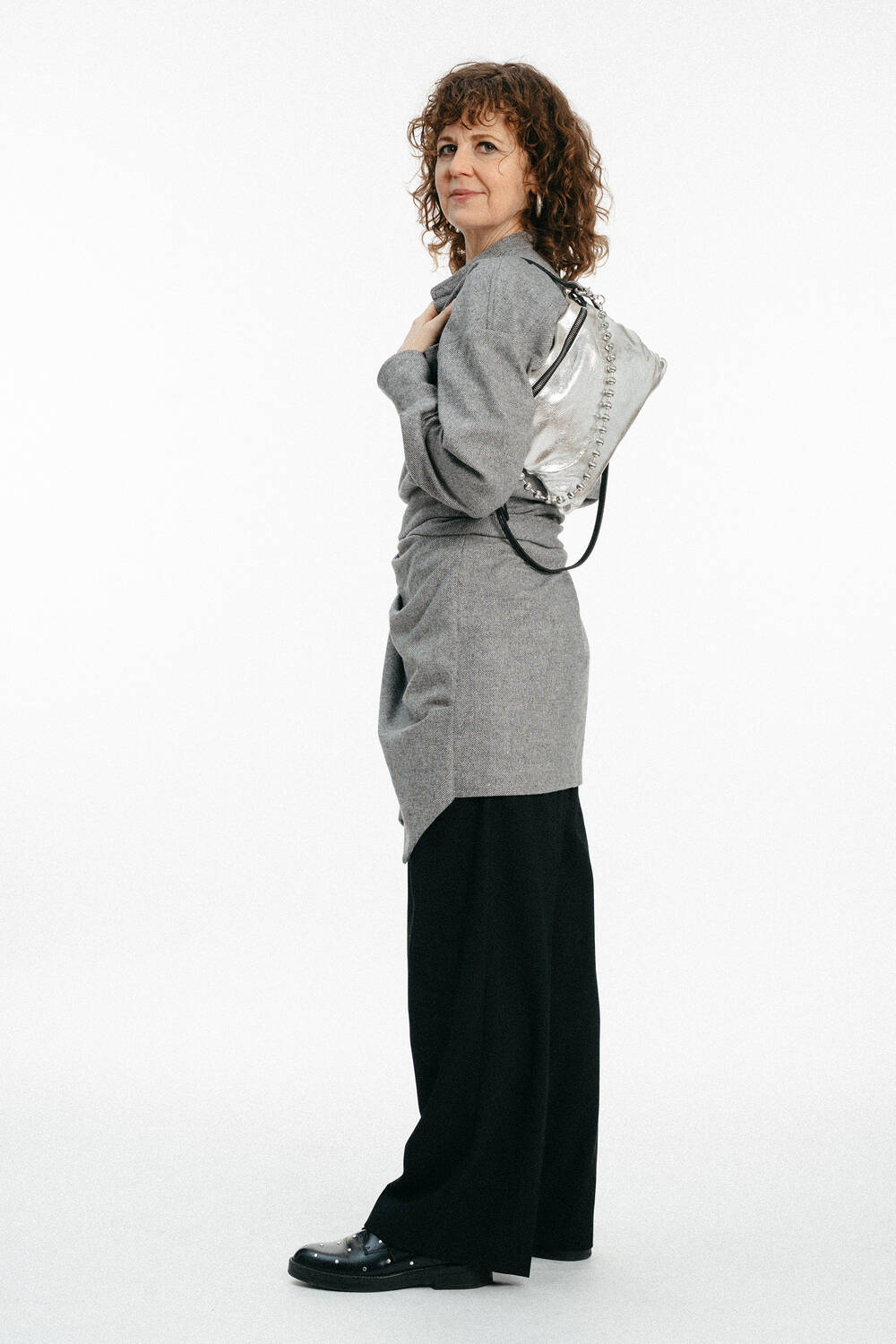 Eine Frau mit lockigem Haar steht seitlich und trägt ein graues langärmeliges Oberteil, eine schwarze Hose mit weitem Bein, schwarze Schuhe und einen silberfarbenen INA KENT-Rucksack über einer Schulter.