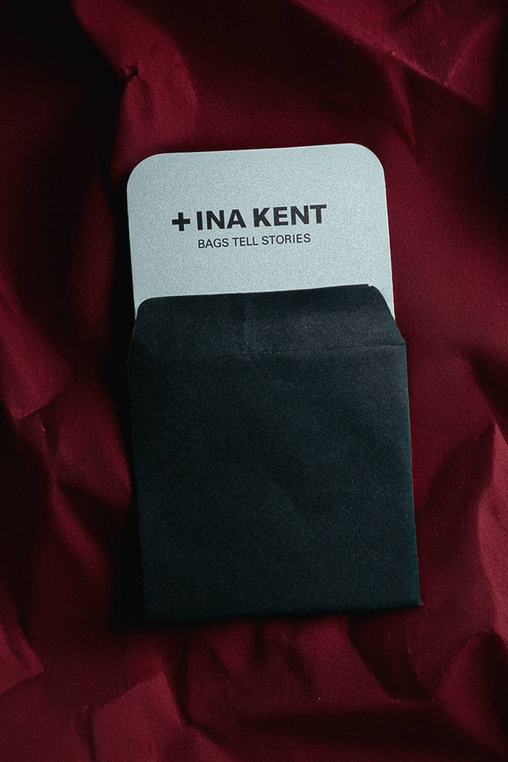 A black envelope partially reveals an INA KENT card with the text "+INA KENT BAGS TELL STORIES," set against a dark red, textured background.