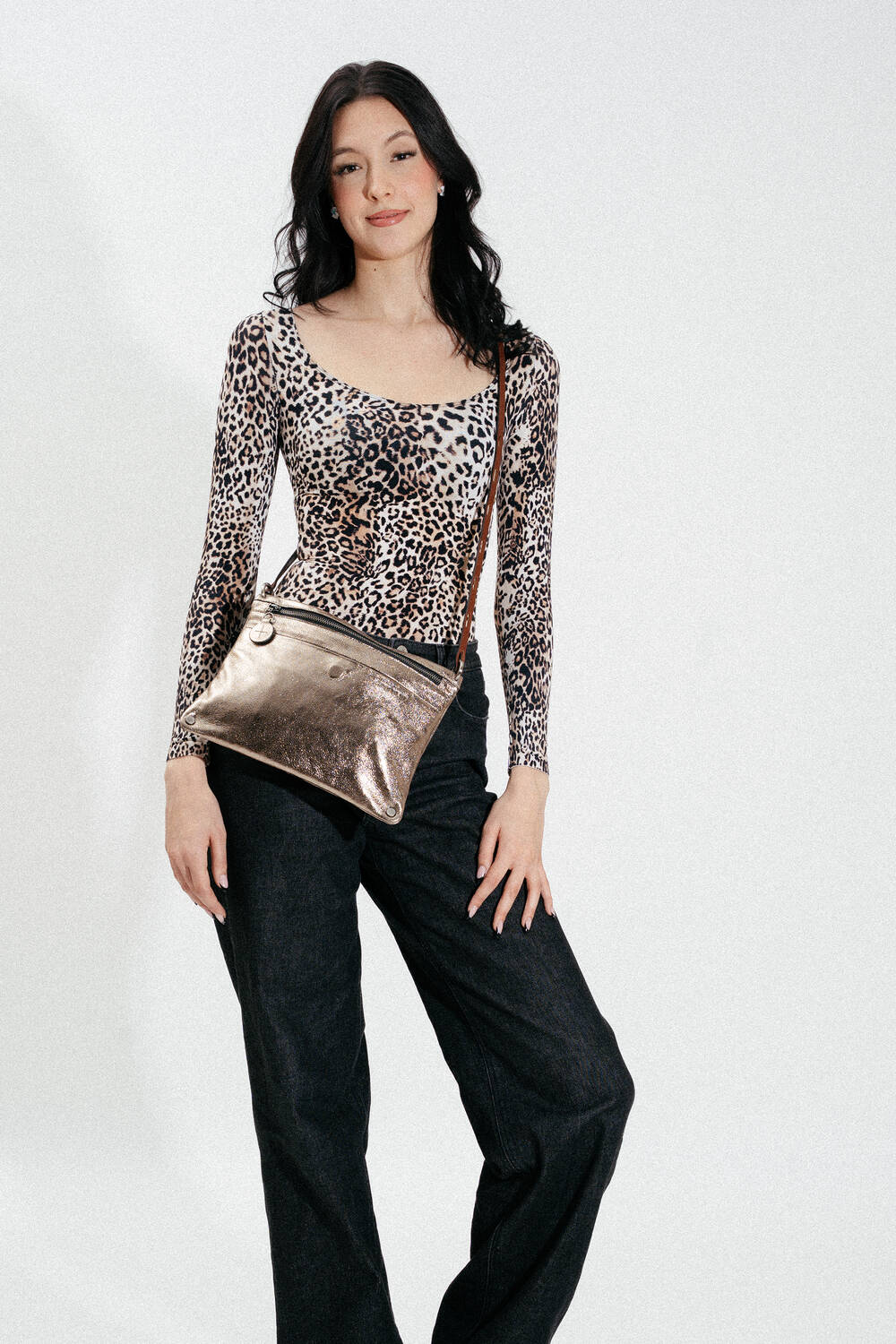 Eine Frau, die ein Oberteil mit Leopardenmuster und eine dunkle Hose trägt und eine INA KENT Metallic-Crossbody-Tasche mit sich führt, steht vor einem schlichten weißen Hintergrund.