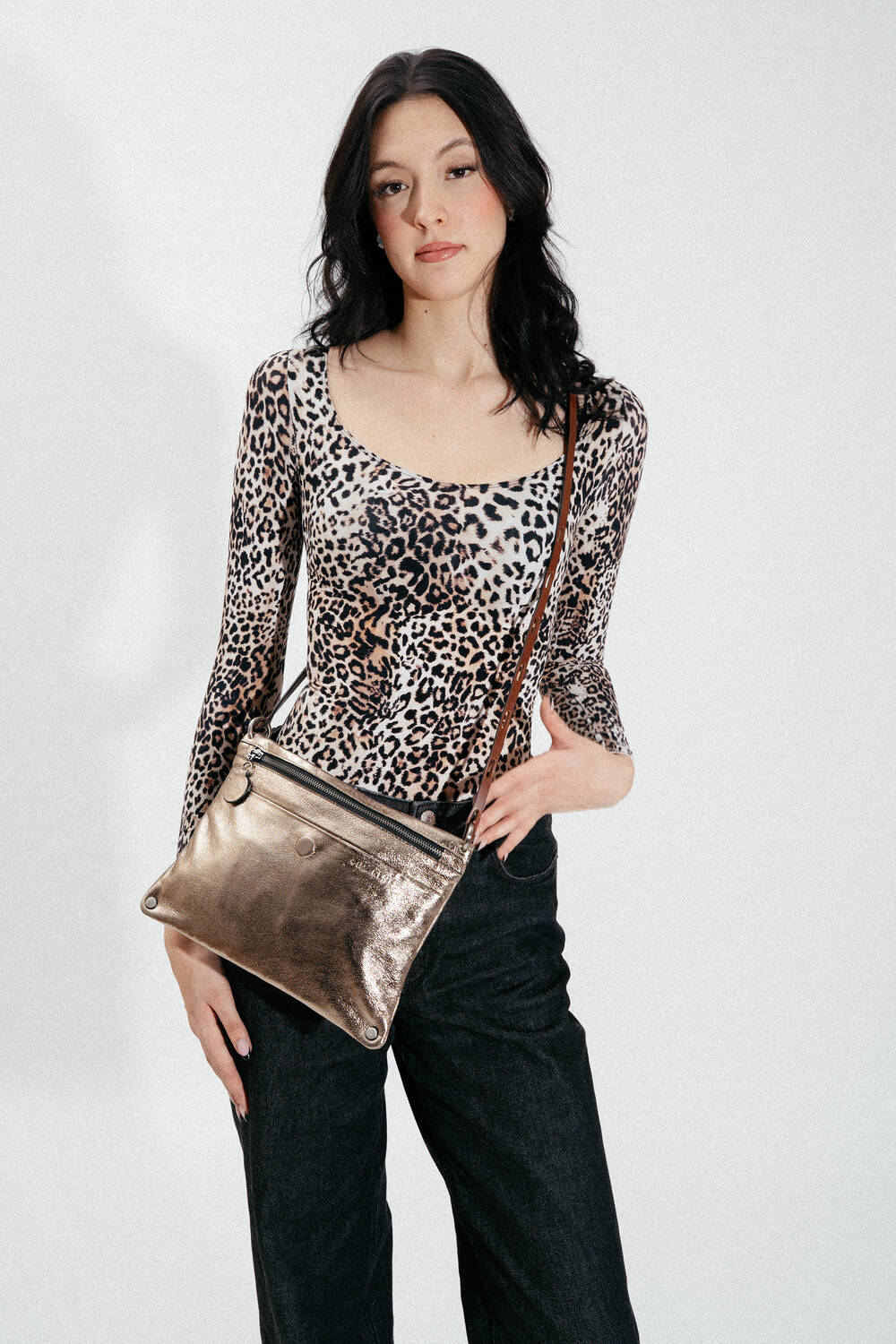 Eine Frau in einem Oberteil mit Leopardenmuster und einer schwarzen Hose hält eine INA KENT Metallic-Crossbody-Tasche vor einem schlichten weißen Hintergrund.