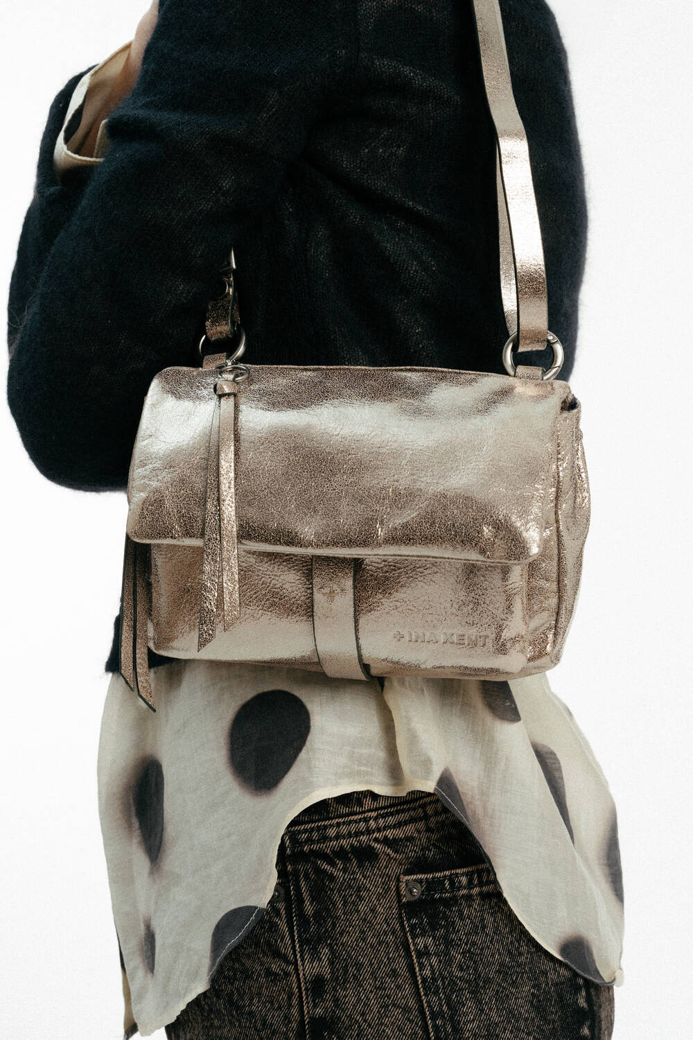 A person wearing a black jacket and printed blouse carries an INA KENT metallic gold crossbody bag with a tassel zipper detail.
