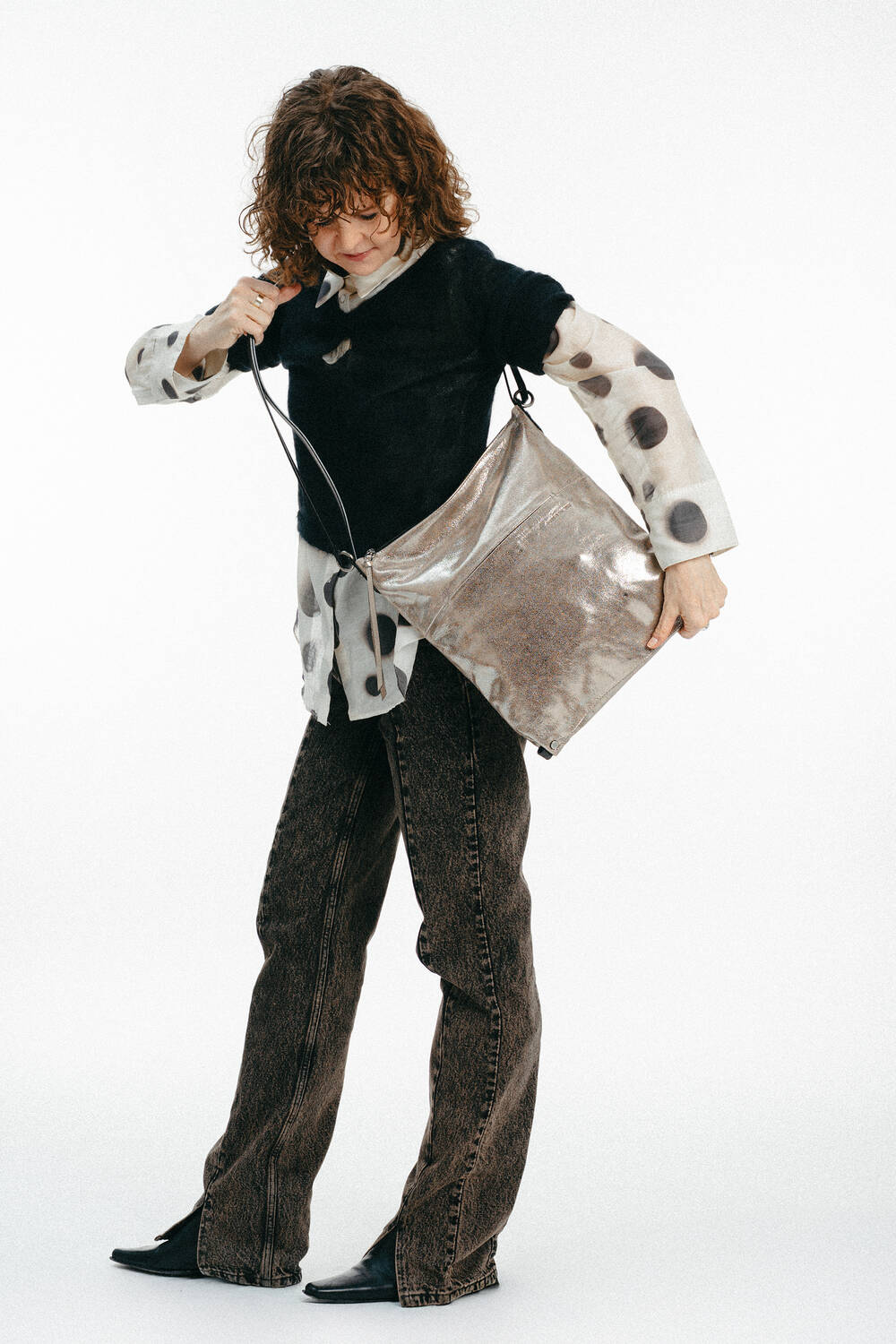 Person, die ein schwarzes Oberteil über einem durchsichtigen gepunkteten Hemd, braune Jeans und schwarze Schuhe trägt und in eine große metallene INA KENT-Umhängetasche schaut.