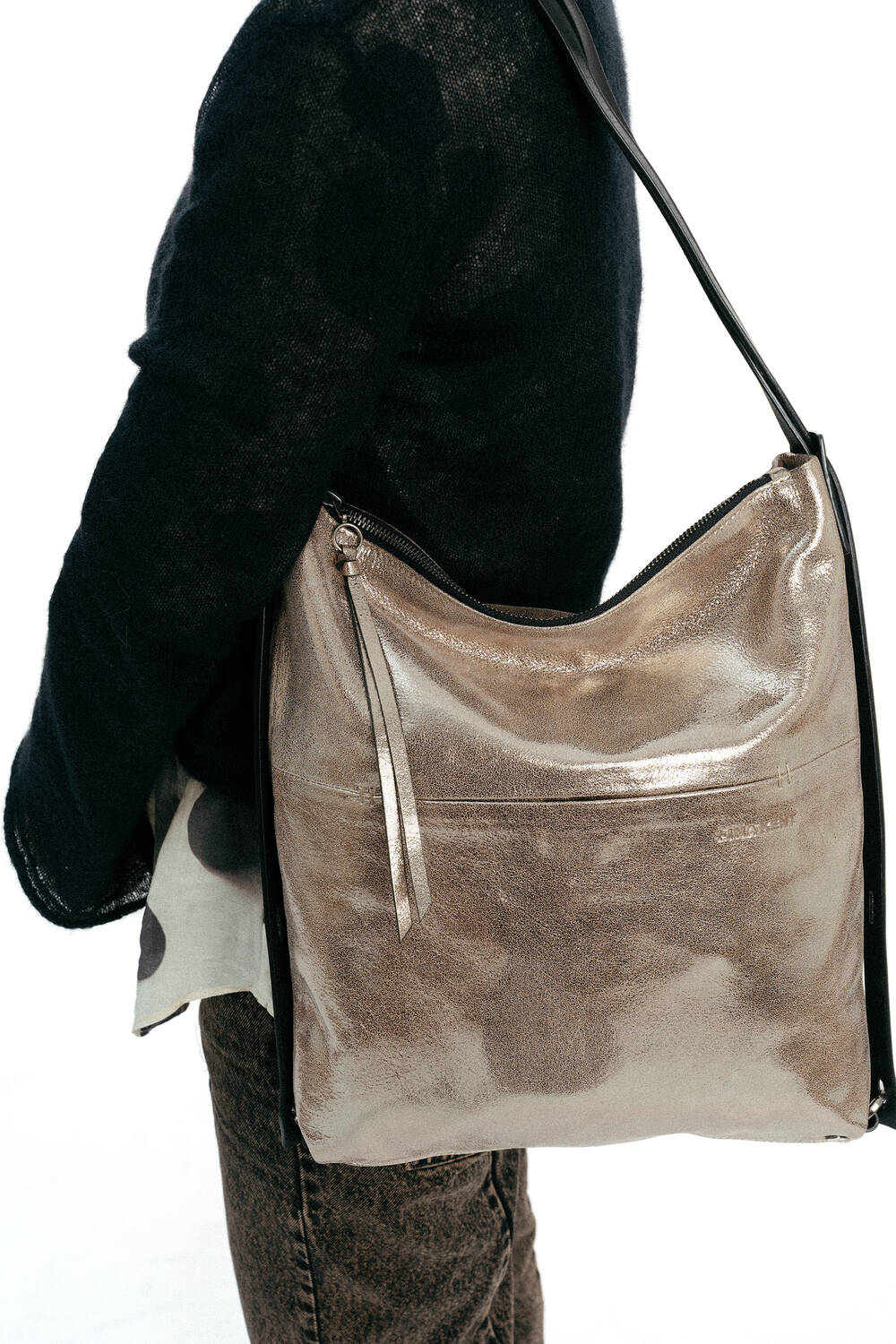 Eine Person, die einen schwarzen Pullover und eine braune Hose trägt, hat eine große INA KENT-Umhängetasche in Goldmetallic mit Reißverschluss und langem Riemen dabei.
