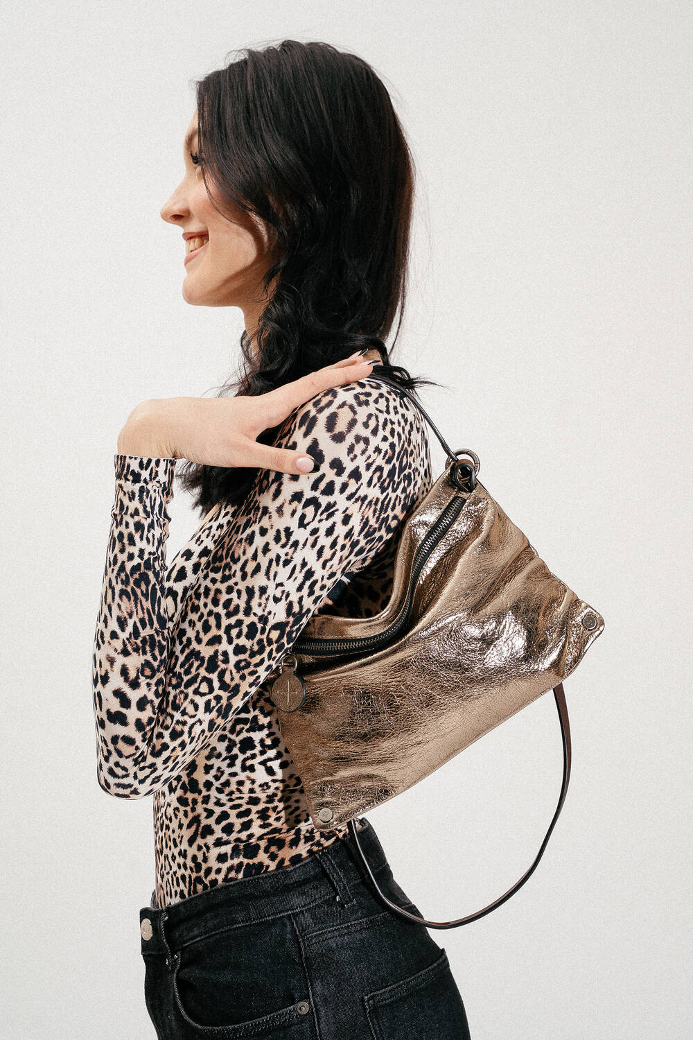 Woman in a leopard print top and black jeans standing sideways with an INA KENT metallic shoulder bag resting on her shoulder.