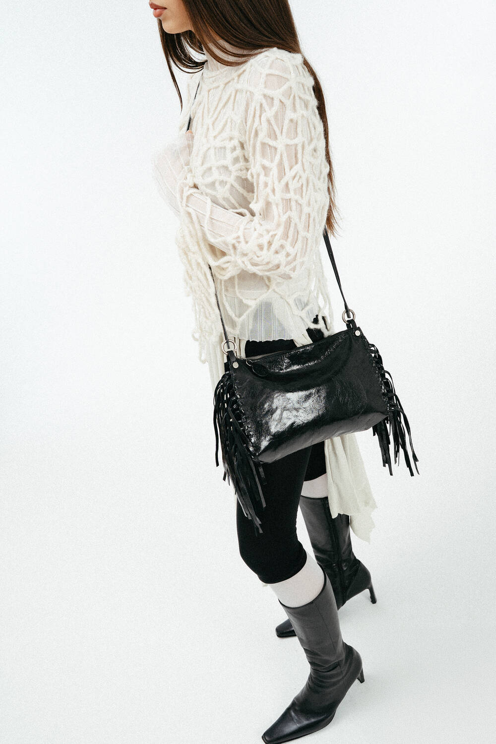 Weiblich gelesenes Model in losem, weißen Strick, mit schwarzer Ledertasche mit Fransen Crossbody getragen
