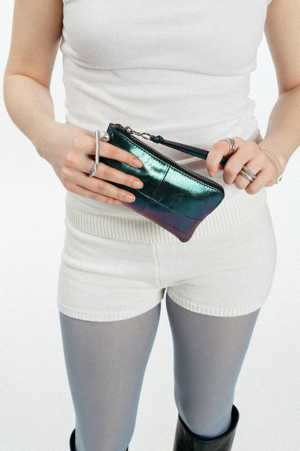 Eine Person, die ein weißes Tanktop, weiße Shorts und eine hellgraue Strumpfhose trägt, hält mit beiden Händen eine kleine metallene INA KENT-Reißverschlusstasche.