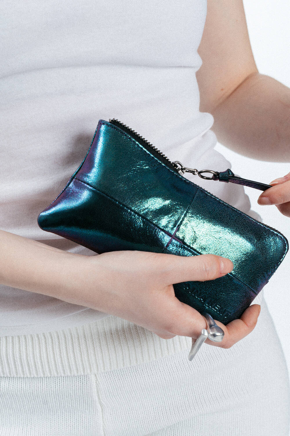 Person mit einer glänzenden, metallisch blauen INA KENT-Clutch-Tasche mit Reißverschluss und einem Schlüssel mit einem herzförmigen Schlüsselanhänger, bekleidet mit einem weißen ärmellosen Oberteil und einer weißen Hose.