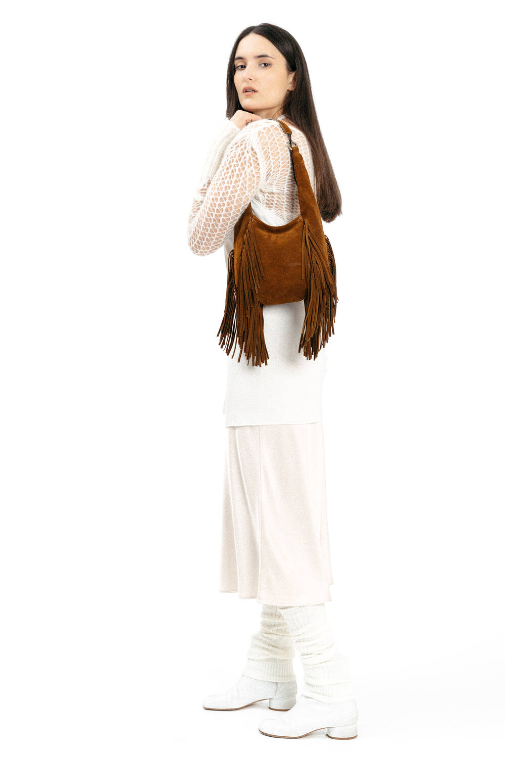 A woman in a white outfit and boots holds an INA KENT brown fringed shoulder bag, standing against a white background.