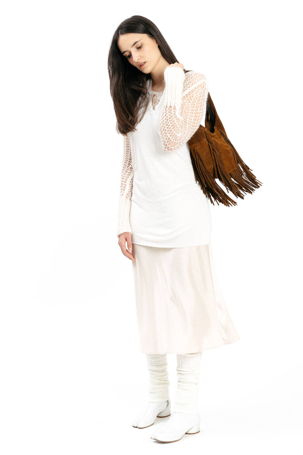 Woman standing against a white background, wearing a long white dress and white boots, carrying an INA KENT brown fringed bag over her shoulder.
