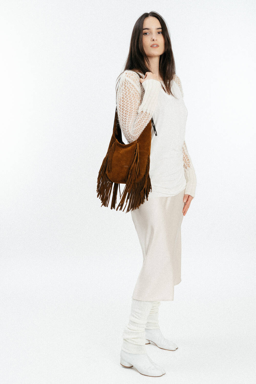 Woman standing against a white background, wearing a cream sweater, light skirt, white boots, and carrying an INA KENT brown fringed shoulder bag.