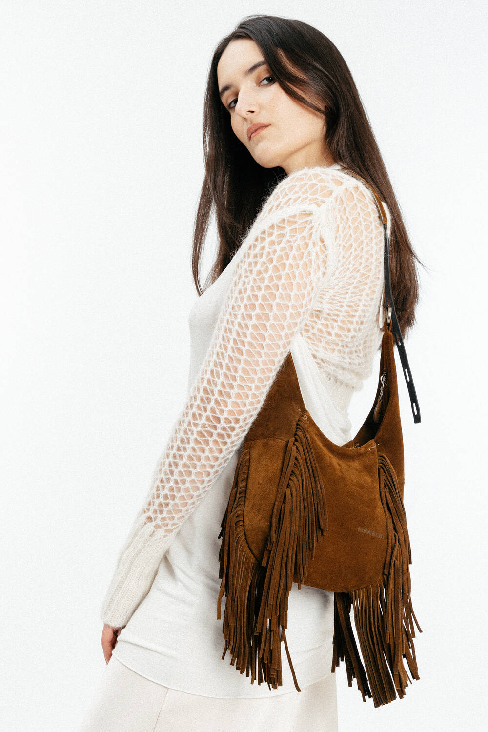 A woman wearing a white knit sweater and skirt poses with an INA KENT brown suede fringe bag over her shoulder against a plain white background.