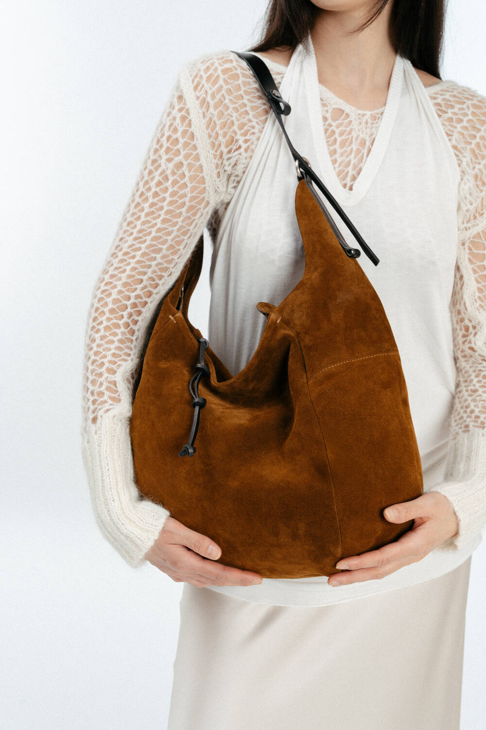A woman wearing a white lace-sleeve top and skirt holds a large brown INA KENT suede shoulder bag with a black strap.