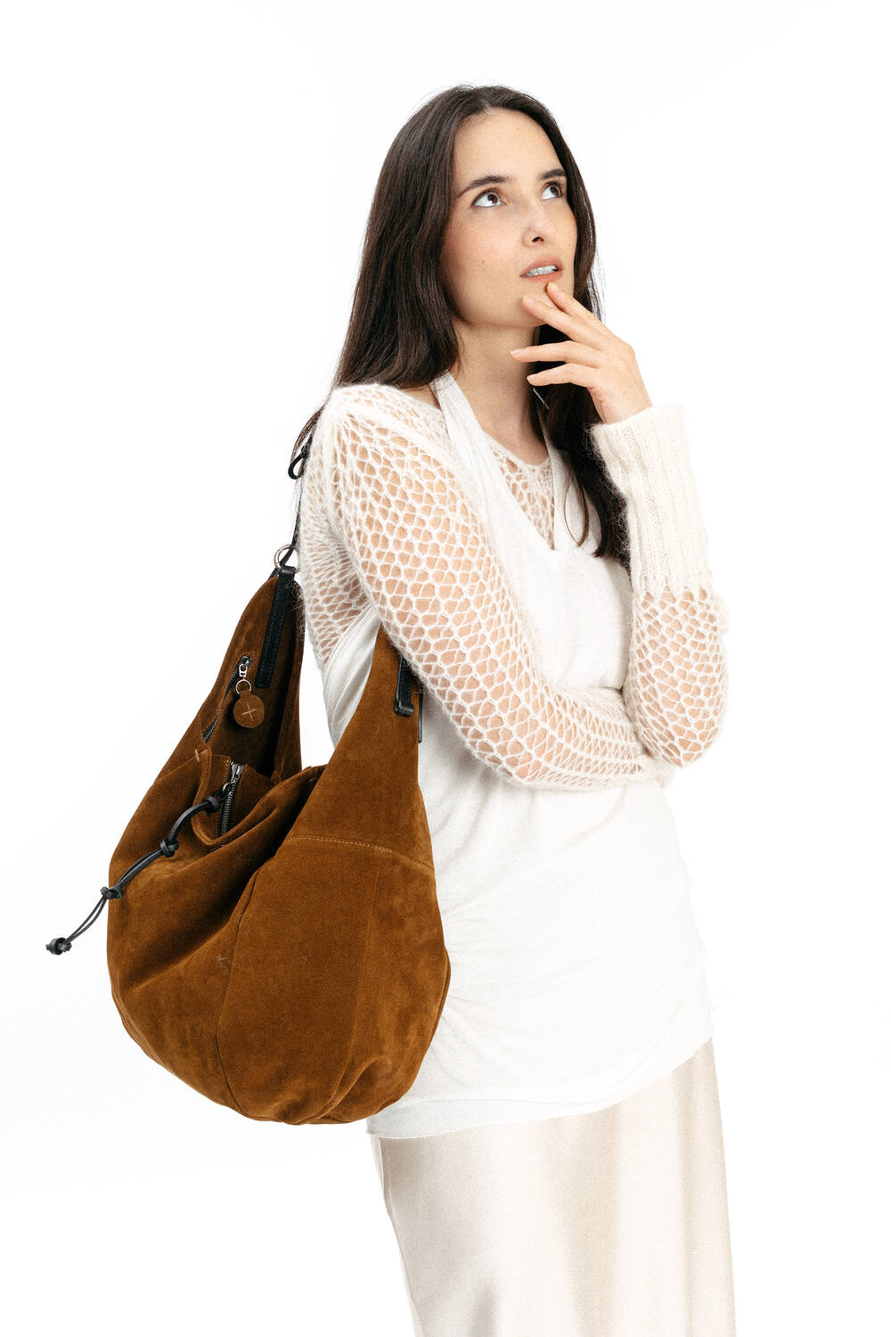 Woman with long dark hair wearing a white textured sweater and cream skirt, carrying a large brown suede INA KENT shoulder bag, standing with one hand on her chin against a white background.