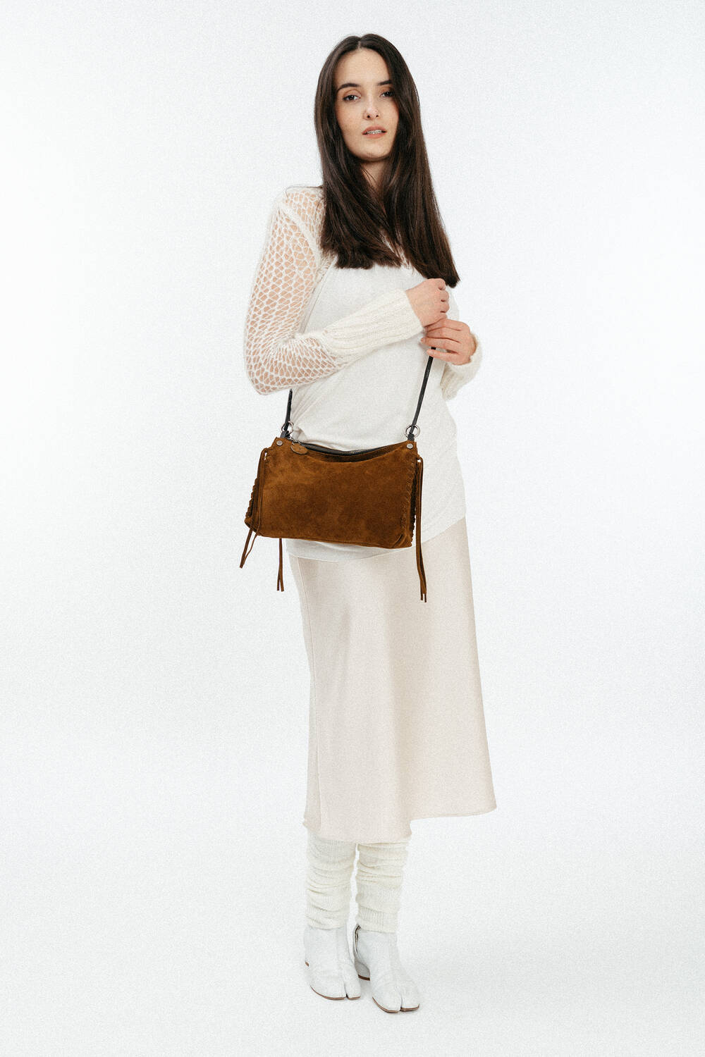 A woman wearing a white long-sleeve top, cream skirt, leg warmers, and white shoes stands holding an INA KENT brown suede shoulder bag against a plain white background.