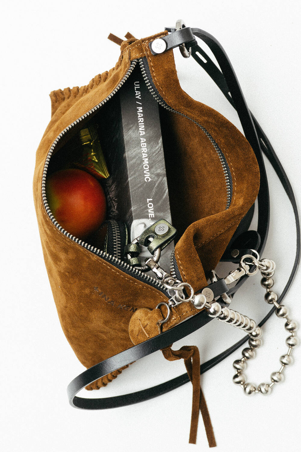 A brown suede INA KENT handbag with a zipper, partially open to reveal a book, a tomato, a lipstick, and a keychain inside, placed on a white surface.