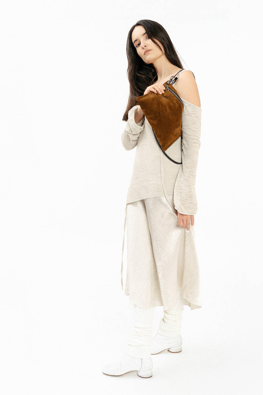 A woman in a light beige outfit and white shoes stands holding an INA KENT brown handbag against a plain white background.