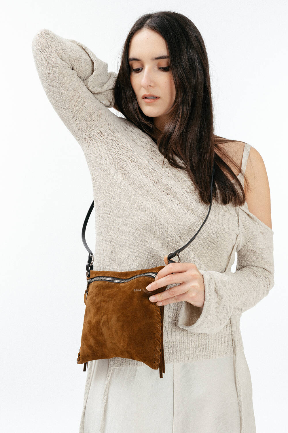 A woman in a light beige off-shoulder sweater holds an INA KENT brown suede crossbody bag with a black strap against a plain white background.
