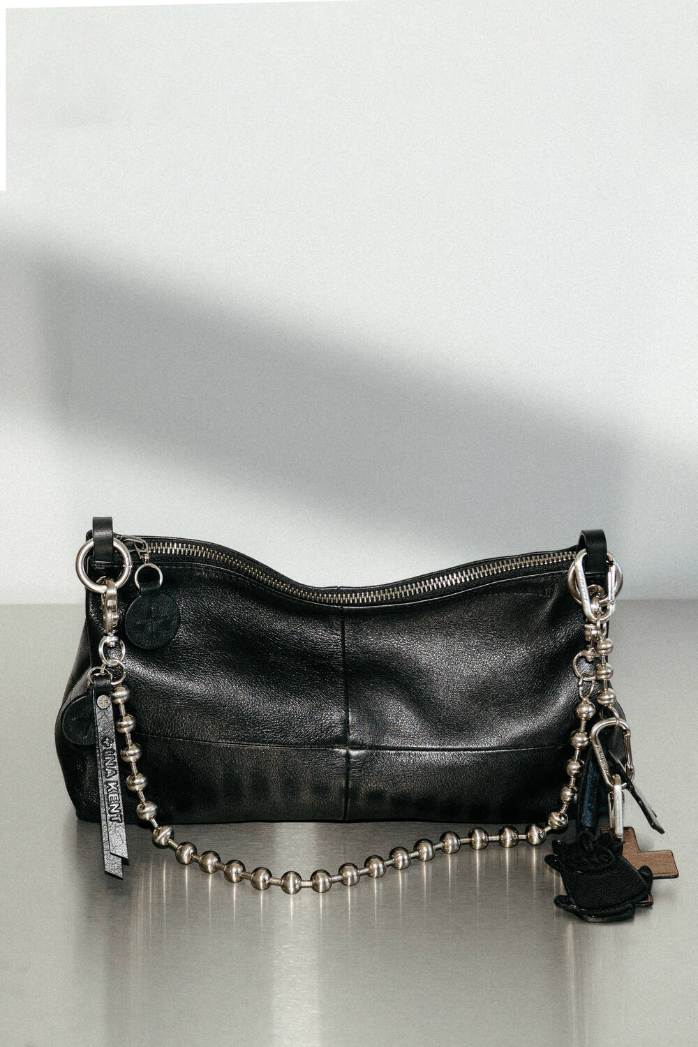 An INA KENT black leather handbag with a silver zipper, metal chain strap, and various keychains and charms attached, placed on a gray surface against a plain background.