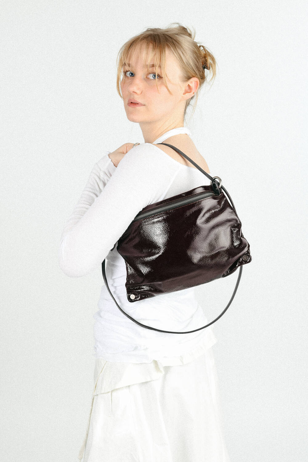 A woman in a white outfit looks over her shoulder, modeling an INA KENT shiny dark brown shoulder bag with a circular handle.