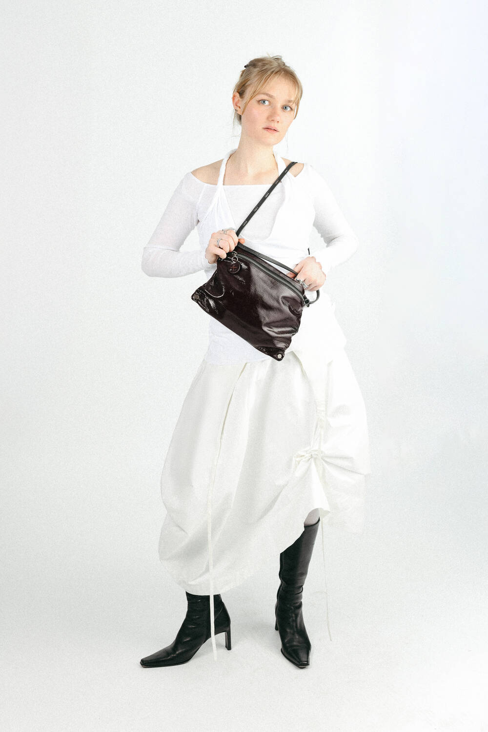 A person in a white long-sleeve top and white skirt stands holding an INA KENT black shoulder bag, wearing black heeled boots against a plain white background.