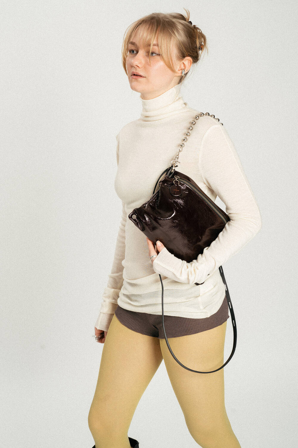 A person wearing a cream turtleneck, brown shorts, yellow tights, and holding a shiny black INA KENT handbag with a chain strap.