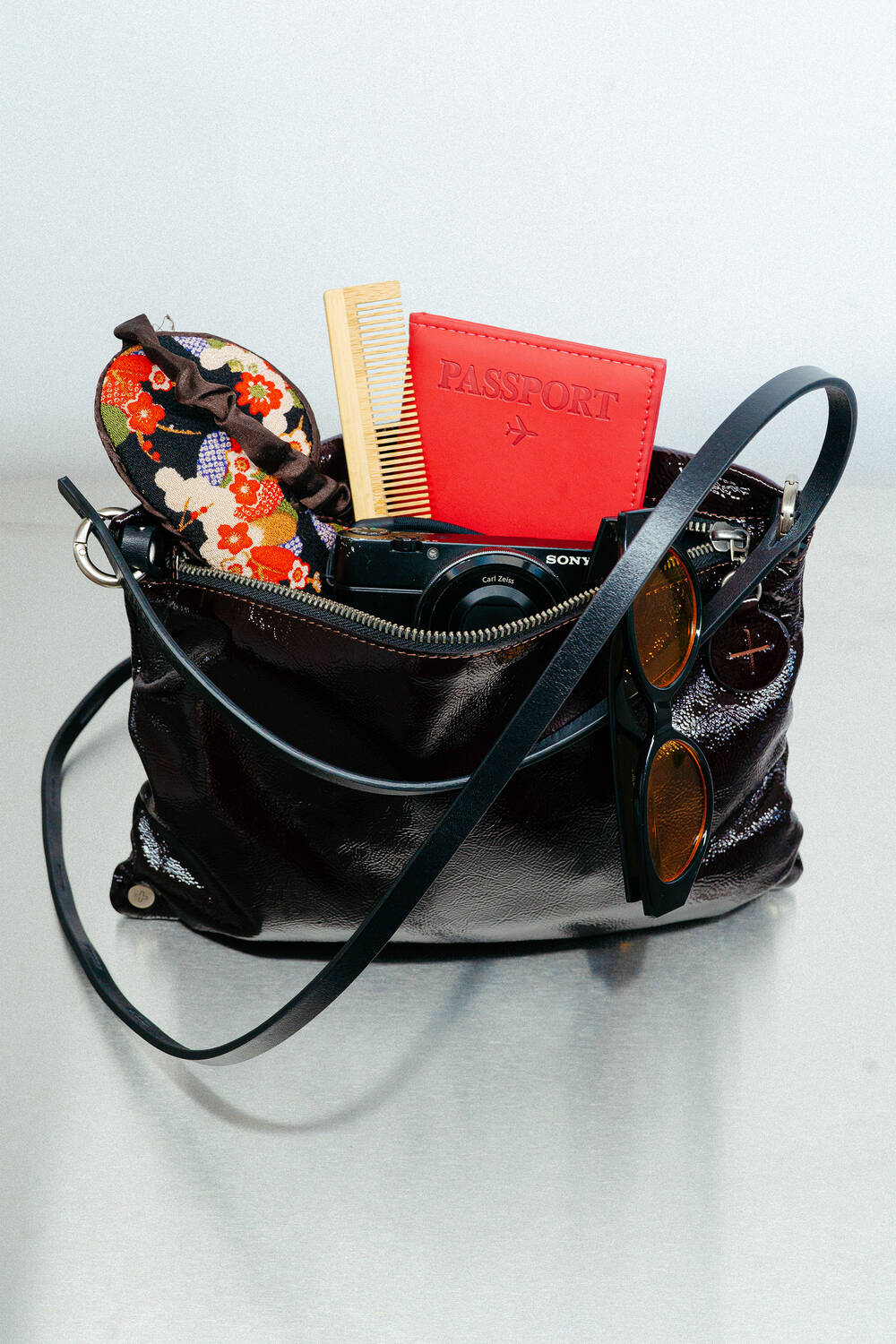 A black INA KENT handbag on a table contains a floral eye mask, a comb, a red passport holder, a camera, and sunglasses.