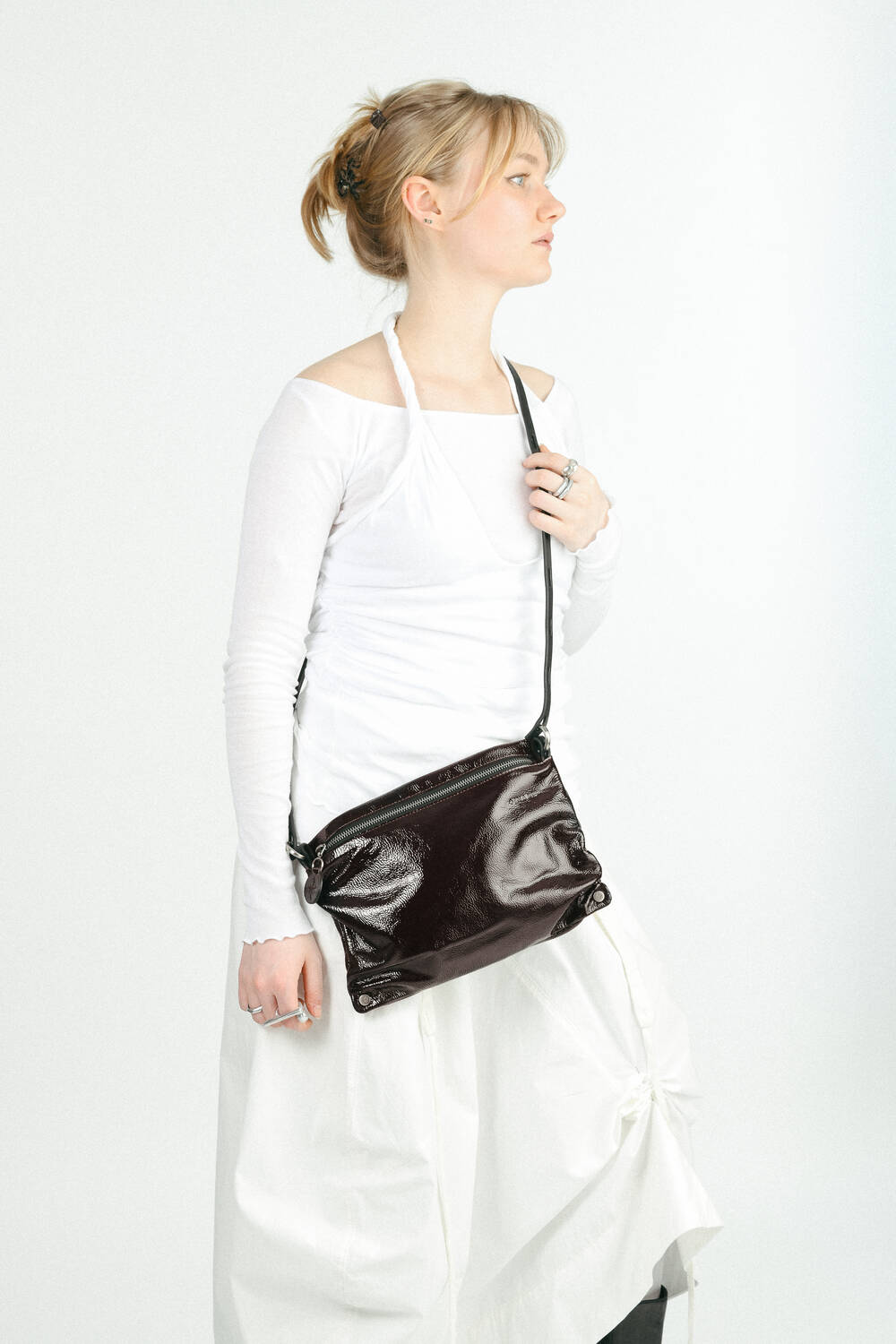 A person wearing a white long-sleeve top and skirt stands against a white background, holding a shiny black INA KENT crossbody bag.