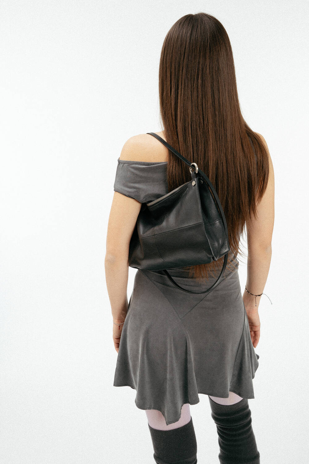 A person with long brown hair, wearing a gray off-the-shoulder dress, black INA KENT bag, white tights, and gray leg warmers stands against a plain white background, facing away.