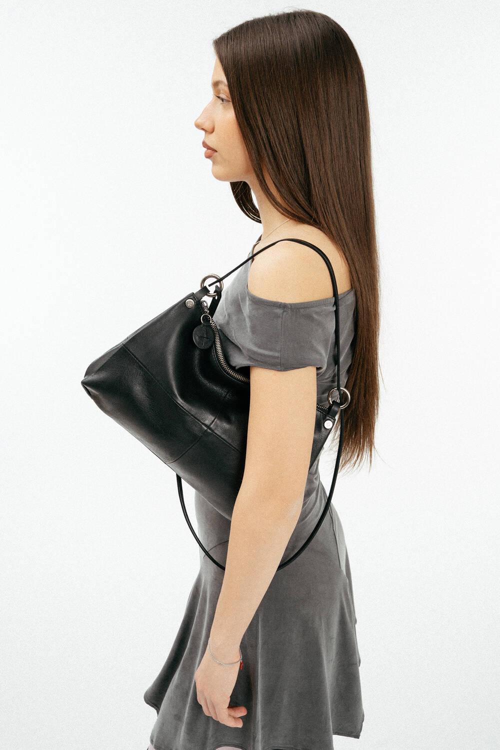 A woman in a grey off-shoulder dress stands in profile, holding an INA KENT black handbag over her shoulder against a plain white background.