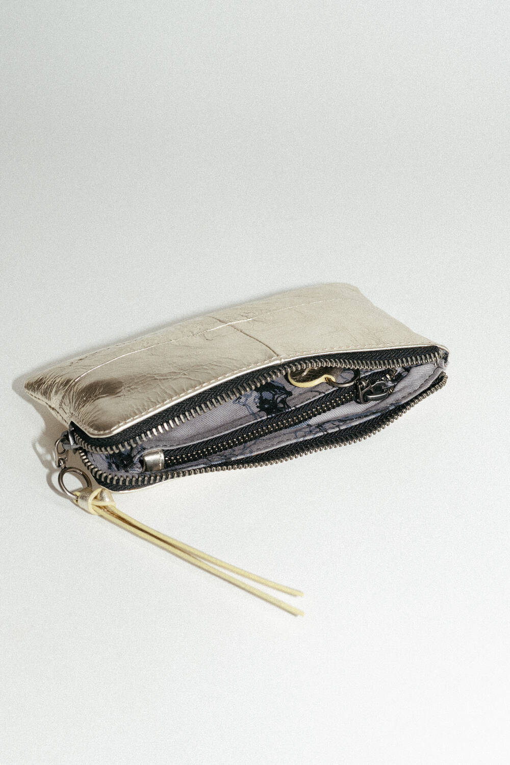 Ein metallischer Beutel mit Reißverschluss von INA KENT ist teilweise geöffnet und gibt den Blick auf ein gemustertes Innenfutter frei. Der Beutel verfügt über einen Reißverschlusszug aus Leder, der mit zwei gelben Quasten verziert ist.