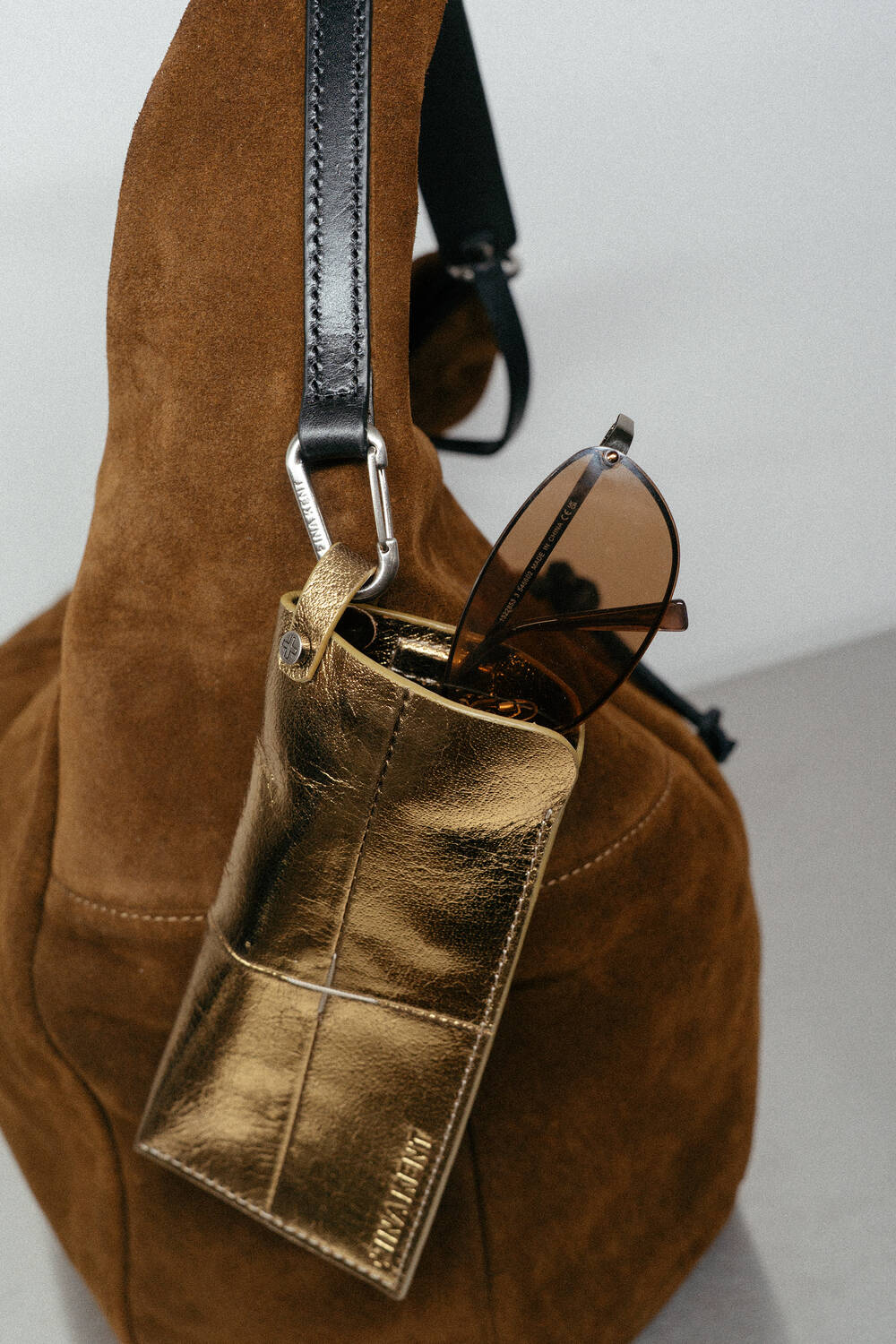 A pair of brown sunglasses is partially tucked into a shiny gold INA KENT leather case, which is attached to the handle of a brown suede bag.