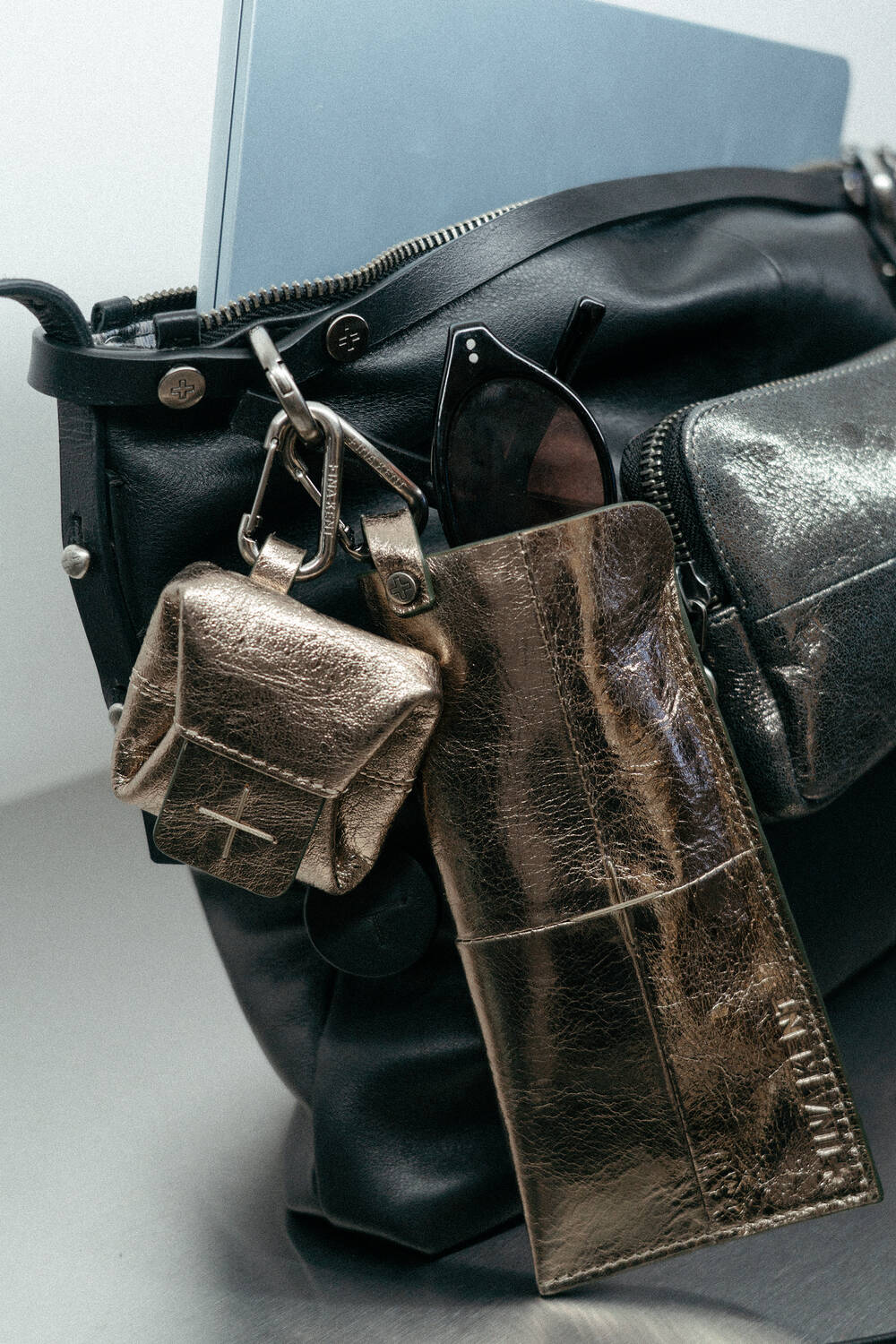 An INA KENT black leather bag with a laptop inside, a metallic gold pouch, sunglasses in a gold case, and a small gold accessory attached to the handle.