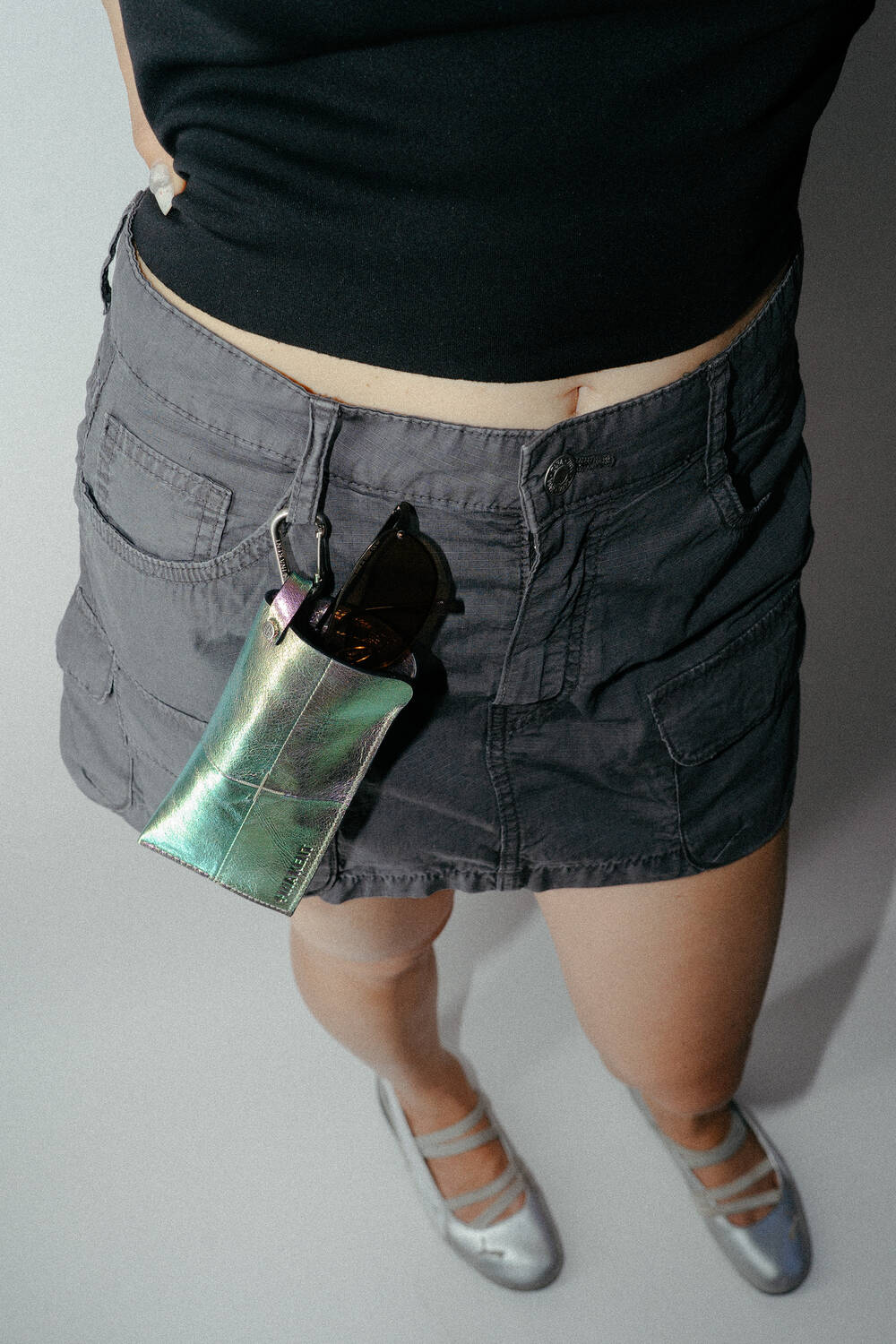 Eine Person, die ein schwarzes Oberteil, einen grauen Cargo-Rock und silberne Schuhe trägt; an der Gürtelschlaufe des Rocks ist eine metallische INA KENT-Tasche befestigt, in der eine Sonnenbrille steckt.