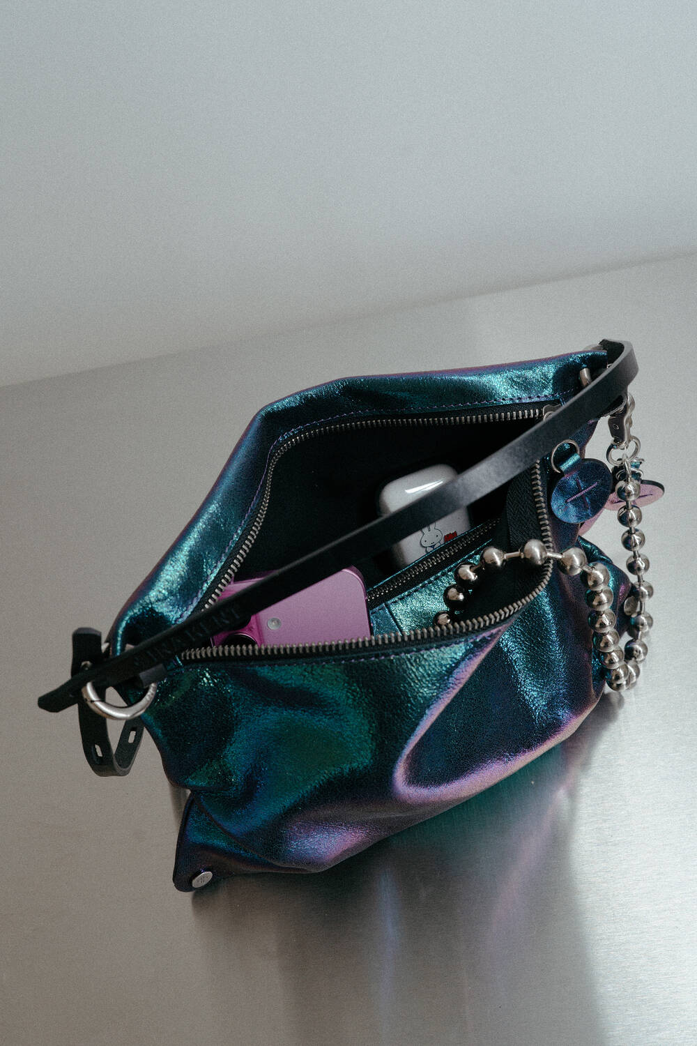 An iridescent INA KENT handbag sits open on a metal surface, containing a phone, a purple card, sunglasses, and a beaded chain keychain.