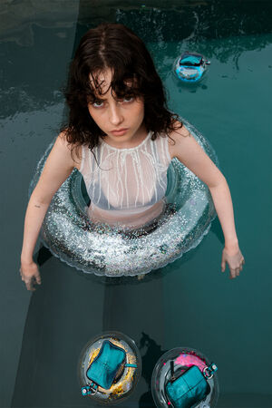 Ein Mädchen mit Anfang zwanzig steht mit einem Luftreifen in einem Pool. Vor ihr schwimmen kleine INA KENT-Portemonnaies X.LOMI ed.2 aus blaugrünem Metallic-Leder