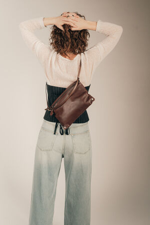 Das Model steht mit dem Rücken zum Betrachter, gekleidet in Jeans, einen hellen Pullover mit einem dunkelblauen Korsett drüber, trägt eine INA KENT Crossbody Tasche MOONLIT ed.1 aus brauen Glattleder