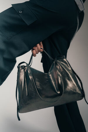 Ein Model, von dem nur die Beine in schwarzer Hose gekleidet, zu sehen sind, hält eine INA KENT Handtasche ROVE ed.1 aus Metallic-Leder in grau zwischen ihren Beinen hindurch.