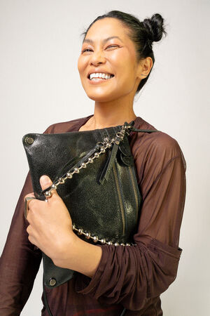 A person with dark hair styled in two buns smiles while holding an INA KENT large black leather bag adorned with a chain detail. The person is wearing a sheer brown top.