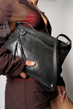 A person in a sheer brown top and skirt holds an INA KENT black textured handbag with a zipper and adjustable strap.