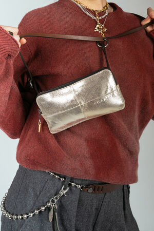 A person wearing a red sweater and gray pants holds an INA KENT metallic crossbody bag across their chest. They have layered necklaces and a chain accessory attached to their pants.