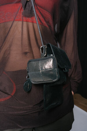 A person wearing a sheer, dark-colored shirt with a small, black INA KENT crossbody bag strapped across their chest. The bag has two compartments and a leather texture.