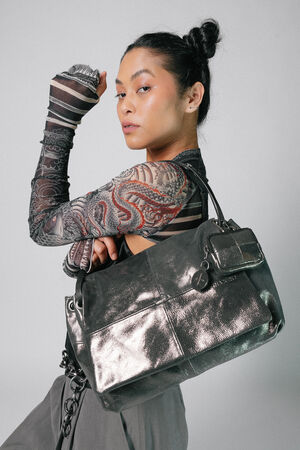 Person with tattoos and a bun hairstyle poses with a large, black INA KENT bag, dressed in a patterned top and gray pants.