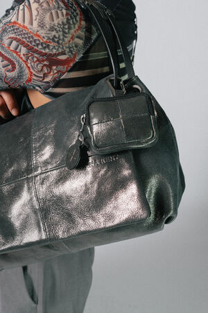Close-up photo of a person holding an INA KENT bag, showcasing its large, metallic leather exterior and a small attached pouch. The person is wearing a long-sleeve shirt with a colorful pattern.