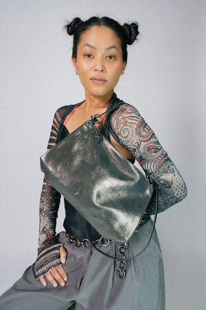 Eine Person mit geflochtenen Haaren und Tattoos trägt ein durchsichtiges langärmliges Oberteil und graue Hosen, hält eine metallische Handtasche von INA KENT mit Kettendetails und posiert vor einem schlichten Hintergrund.