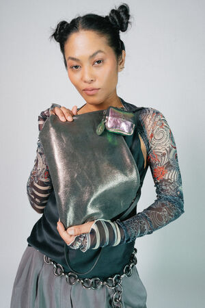 Person with dark hair in two buns holds a metallic INA KENT bag. They wear a sleeveless black top and patterned sleeves, with a small metallic pouch attached to the bag.
