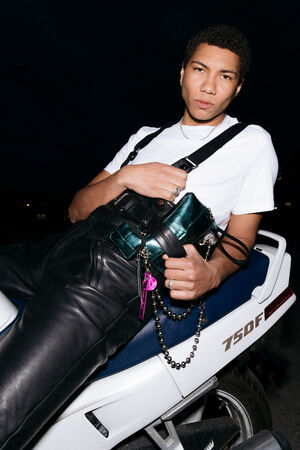 A person with short hair wearing a white shirt and black overalls, sitting on a motorcycle, holding an INA KENT metallic bag with various keychains, against a dark background.