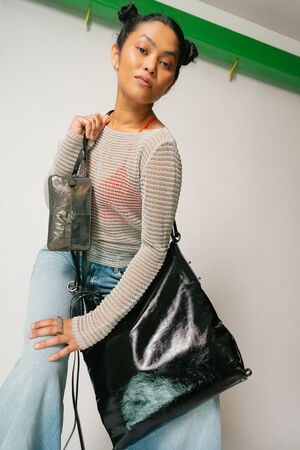 Eine Frau posiert mit einer großen schwarzen Handtasche von INA KENT und einer kleineren metallischen Tasche. Sie trägt ein transparentes Strickoberteil über einem roten BH und hellblauen Jeans. Ihr Haar ist zu zwei Dutts frisiert.