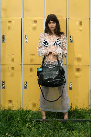 A woman stands in front of yellow lockers, holding a stylish INA KENT black bag. She wears a white netted top over a black outfit and a transparent skirt. Grass surrounds her feet.